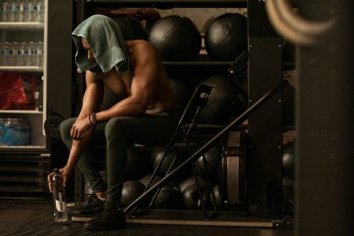 Shirtless man with towel on head seated in gym near weight rack, holding water bottle. Fitness and gym lifestyle shot, low-key.