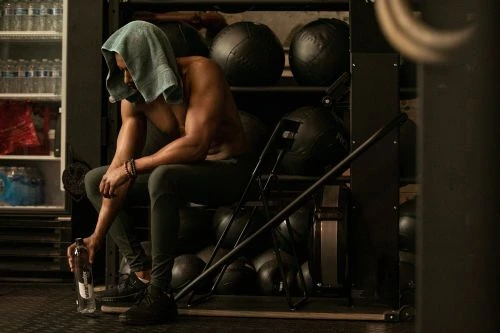 Shirtless man with towel on head seated in gym near weight rack, holding water bottle. Fitness and gym lifestyle shot, low-key.