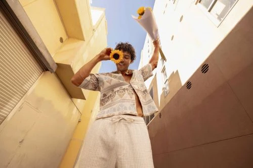 Young adult poses with sunflower in urban setting. Low-angle view shows patterned top, light pants, and surrounding city buildings against blue sky.