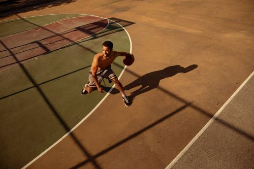 Shirtless adult male athlete dribbling a basketball on an outdoor sports court with visible tree shadows. View from above, dynamic action.