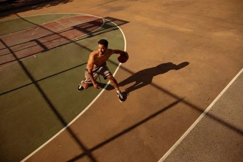 Shirtless adult male athlete dribbling a basketball on an outdoor sports court with visible tree shadows. View from above, dynamic action.