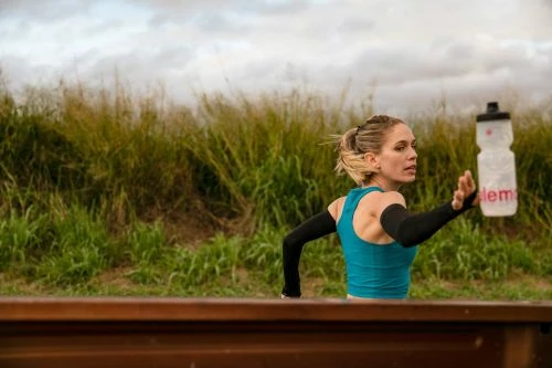 Active woman runs outdoors holding water bottle on a cloudy day in nature, wearing fitness apparel, with green grasses in background.