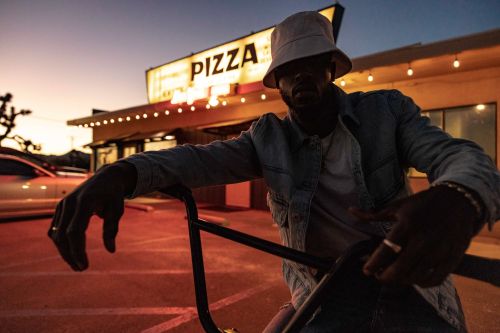Young man in bucket hat and denim jacket poses with bicycle in front of vintage pizza restaurant at twilight. Exterior shot.