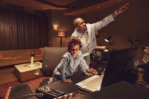 African American man and child in a music studio with sound equipment, computer, mixing board, and studio lighting in a warm environment.