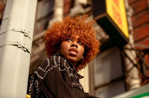 Close-up portrait of a Black woman with voluminous ginger afro curls wearing a dark studded jacket, captured by Andre L. Perry.