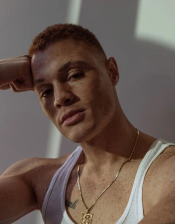 Close-up portrait of a young man with red hair wearing a white tank top and gold chain necklace against a light wall with shadows.