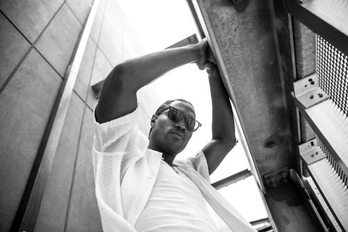 Black and white portrait of a man in sunglasses looking upwards while bracing himself on metal beams with a textured wall behind.