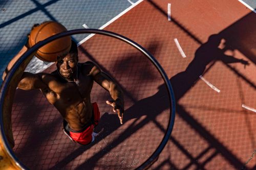 Top-down view of an athletic Black man, shirtless, holding a basketball on an outdoor court, focused through a basketball hoop.