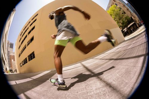 Man in athletic wear running on concrete sidewalk between buildings in an urban setting, captured with fisheye lens and a low-angle perspective.