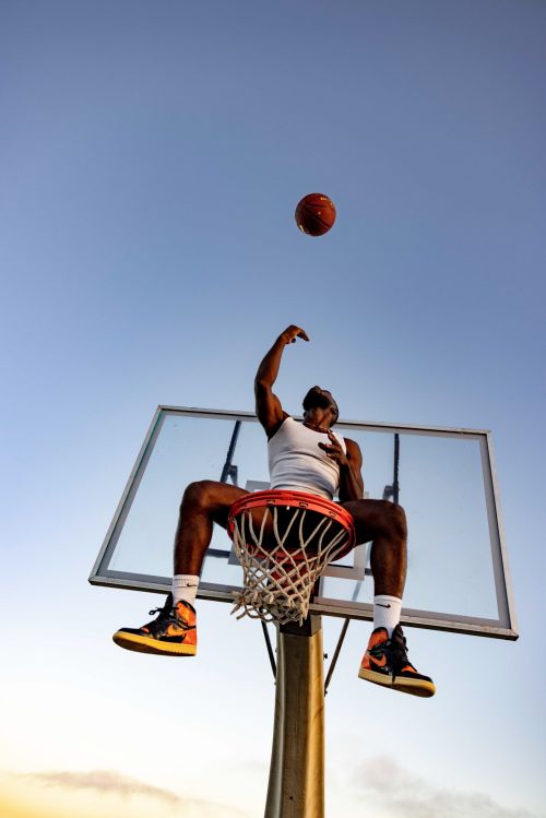 An athletic adult male basketball player sitting on a basketball hoop about to throw a ball into a clear blue sky.