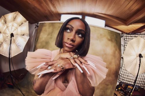 Woman with long white nails poses in a pink dress in a photo studio with umbrellas and a beige backdrop, portrait orientation.