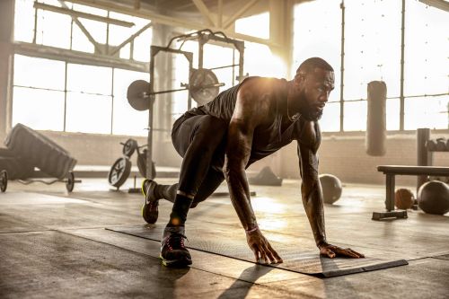 Athletic, muscular man in sportswear does a plank on a mat in a gym setting, with natural light streaming through large windows.