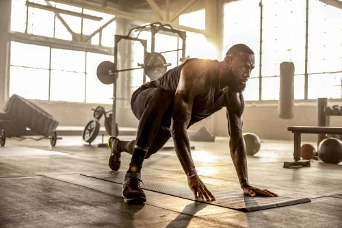 Athletic, muscular man in sportswear does a plank on a mat in a gym setting, with natural light streaming through large windows.