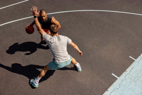 Two adult males playing basketball on an outdoor court. The players are wearing athletic clothing and shoes. The court is grey with white lines.