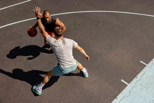 Two adult males playing basketball on an outdoor court. The players are wearing athletic clothing and shoes. The court is grey with white lines.