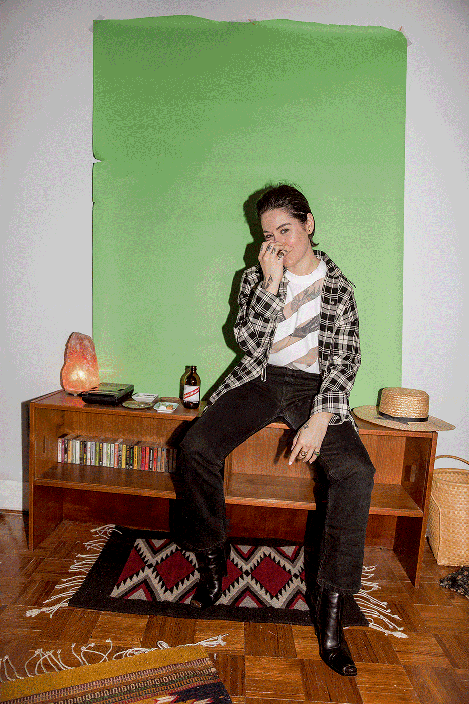 Young adult woman posing on a mid-century modern shelving unit with a green backdrop, illuminated by soft, diffused light in a room.
