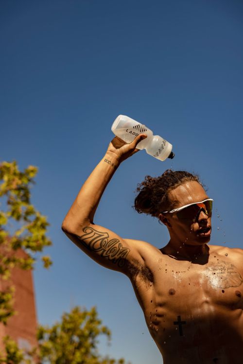 Muscular man outdoors cools off by pouring water from a bottle over himself on a sunny day with blue skies in an urban setting.