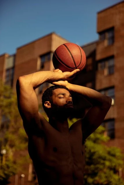 Man with dark skin, shirtless, preparing to shoot a basketball outdoors against the backdrop of a brick apartment building and blue sky.
