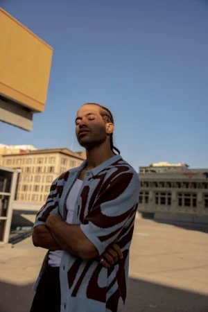 Man with braided hair wearing a patterned blue cardigan standing outdoors under a clear sky in Los Angeles.