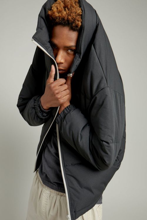 A young adult with curly brown hair, wearing a dark puffer jacket with a white zipper and light pants, stands in a studio environment.