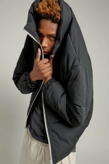 A young adult with curly brown hair, wearing a dark puffer jacket with a white zipper and light pants, stands in a studio environment.