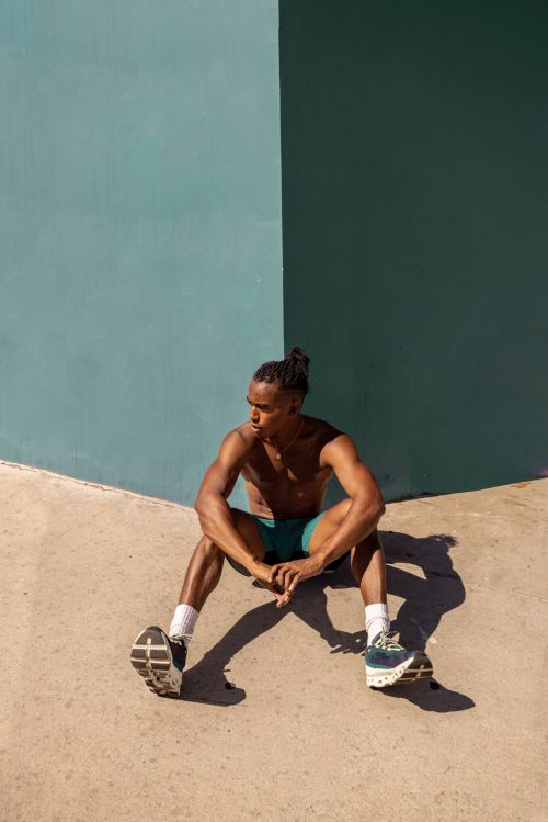 Shirtless black man with braided hair seated against a teal wall in Los Angeles by photographer Andre L. Perry.