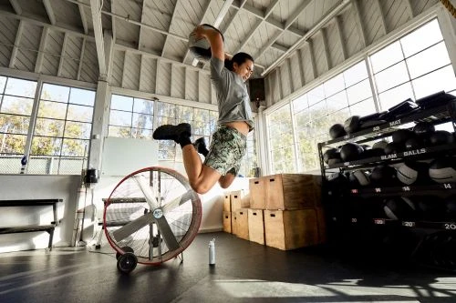 An athletic woman jumps with a slam ball overhead in a bright gym with a large fan, exercise equipment, and natural light from windows.