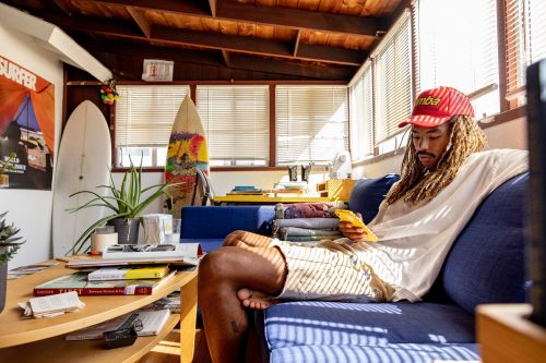 A man sits on a blue sofa in a brightly lit room, looking at a yellow cell phone, with surfboards and plants in the background.