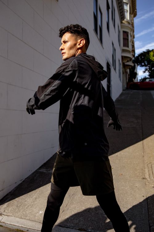Athletic man in black jacket and shorts walks on sidewalk along buildings in San Francisco, California on a sunny day.