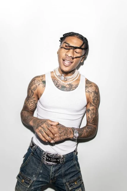 Portrait of a tattooed adult male with braids, wearing a white tank top and denim pants against a white background.