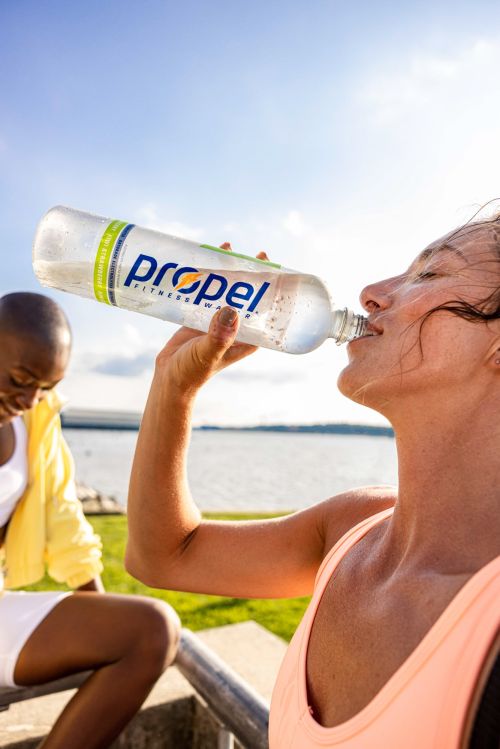 Woman drinking Propel Fitness Water outdoors near lake, another woman sits in background. Clear water bottle features the Propel logo.