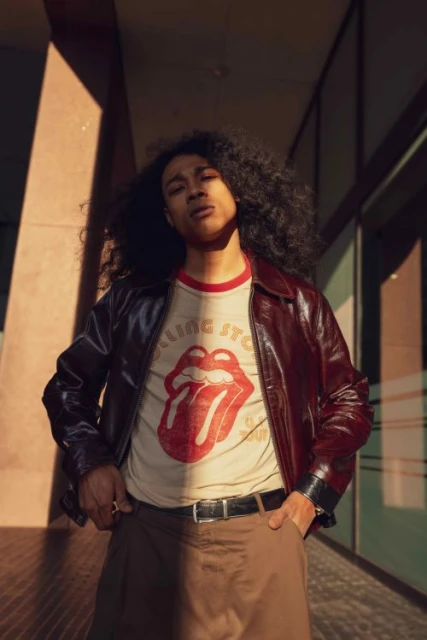 A young adult stands in front of a modern building in brown jacket, t-shirt with the Rolling Stones logo, and curly dark hair.