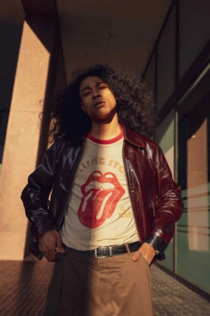 A young adult stands in front of a modern building in brown jacket, t-shirt with the Rolling Stones logo, and curly dark hair.