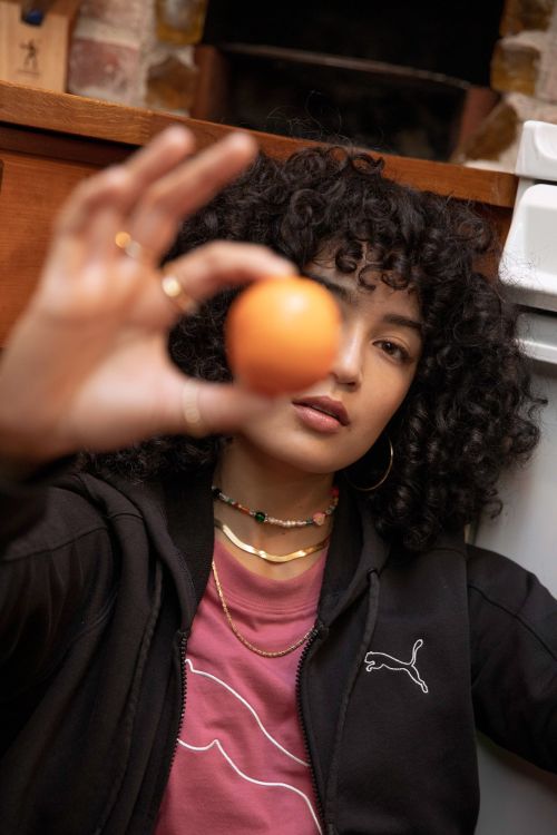A young woman with curly dark hair holds an orange in her hand indoors, wearing a Puma zip-up jacket and gold necklaces.