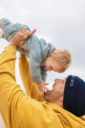 Adult man holding baby in the air, smiling at each other. Man is wearing a yellow sweatshirt and a dark blue beanie outdoors.