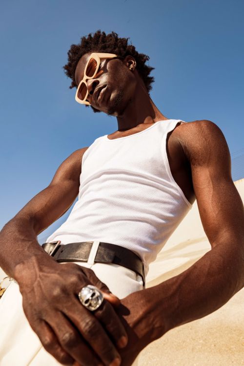 Stylish dark-skinned man in sunglasses, white tank top and pants posing against a bright blue sky background in a low-angle shot.