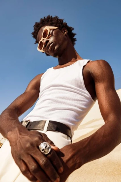 Stylish dark-skinned man in sunglasses, white tank top and pants posing against a bright blue sky background in a low-angle shot.