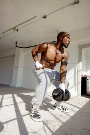 Muscular man in a light-filled room wearing gray sweatpants and lifting a dumbbell while bracing himself, showcasing strength and fitness.