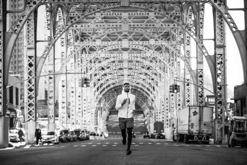 Black and white shot of an African-American man jogging on a city bridge, wearing athletic clothing with vehicles and buildings around.