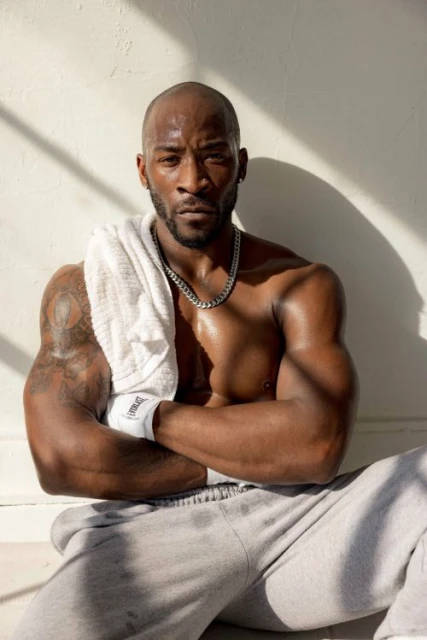 Athletic Black man poses shirtless with sweatpants, towel, and silver chain indoors with sunlight and shadows on a white wall.