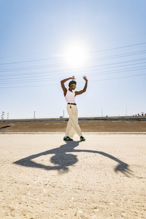 An adult male stands with arms raised in an outdoor, sunlit, desert-like environment. He is wearing sunglasses and light-colored clothing.