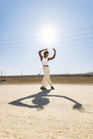 An adult male stands with arms raised in an outdoor, sunlit, desert-like environment. He is wearing sunglasses and light-colored clothing.