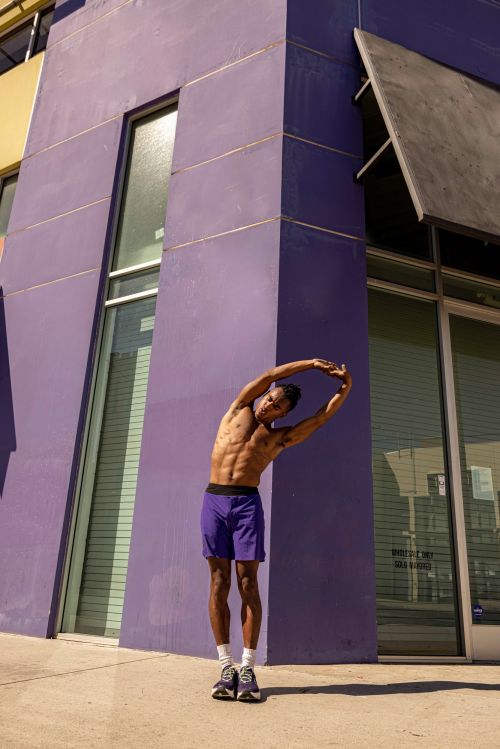 Athletic man with bare torso stretches arms overhead outdoors against a bright purple urban building in full sunlight.