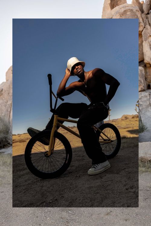 A Black male model with a white bucket hat poses on a yellow bicycle in a desert landscape, framed by a stone-patterned border.