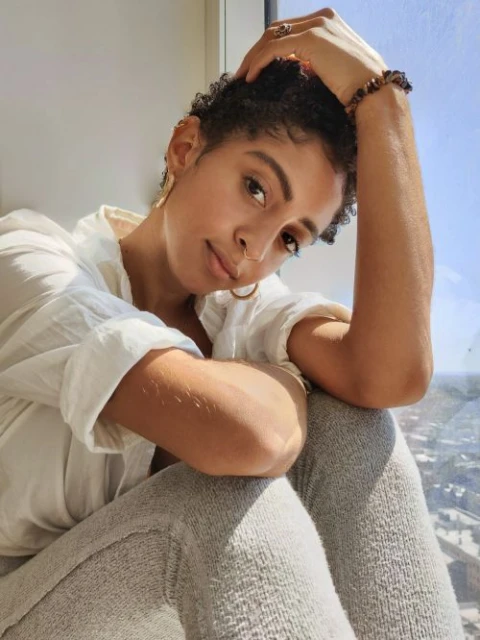 Woman with short curly hair in natural light sits by window in gray leggings and an off-white blouse, accessorized with gold hoop earrings.