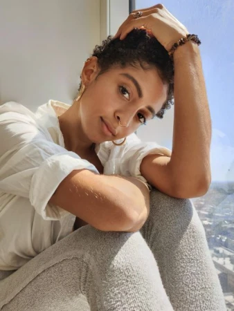Woman with short curly hair in natural light sits by window in gray leggings and an off-white blouse, accessorized with gold hoop earrings.