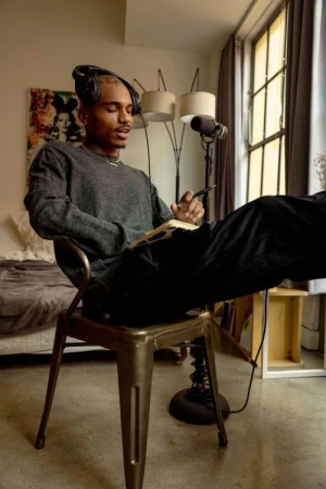Adult male sitting indoors wearing headphones, writing in notebook, microphone, lamp, and large window in the background. 