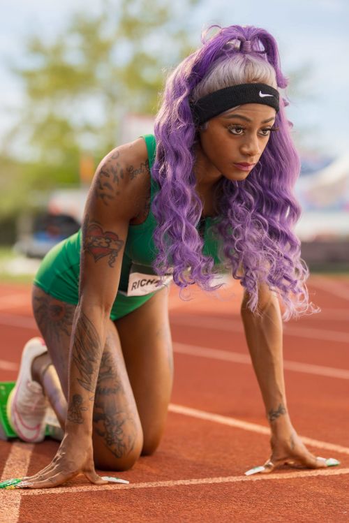 Female athlete with purple hair in starting position on a running track wearing a green uniform and black Nike headband outdoors.