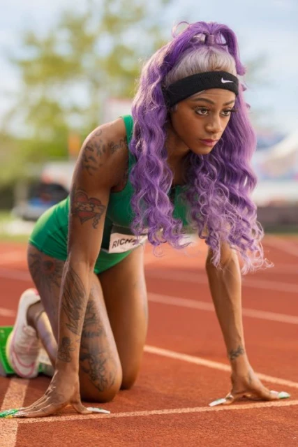 Female athlete with purple hair in starting position on a running track wearing a green uniform and black Nike headband outdoors.