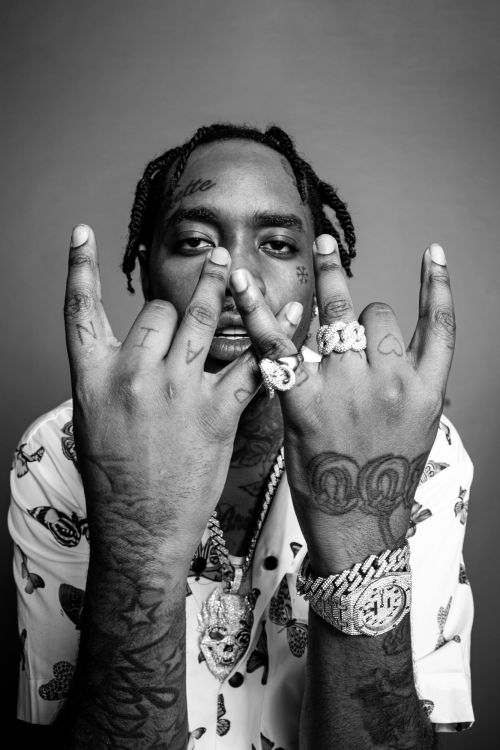 Black and white portrait of a tattooed man with braided hair wearing jewelry and a patterned shirt, making hand gestures in a studio.
